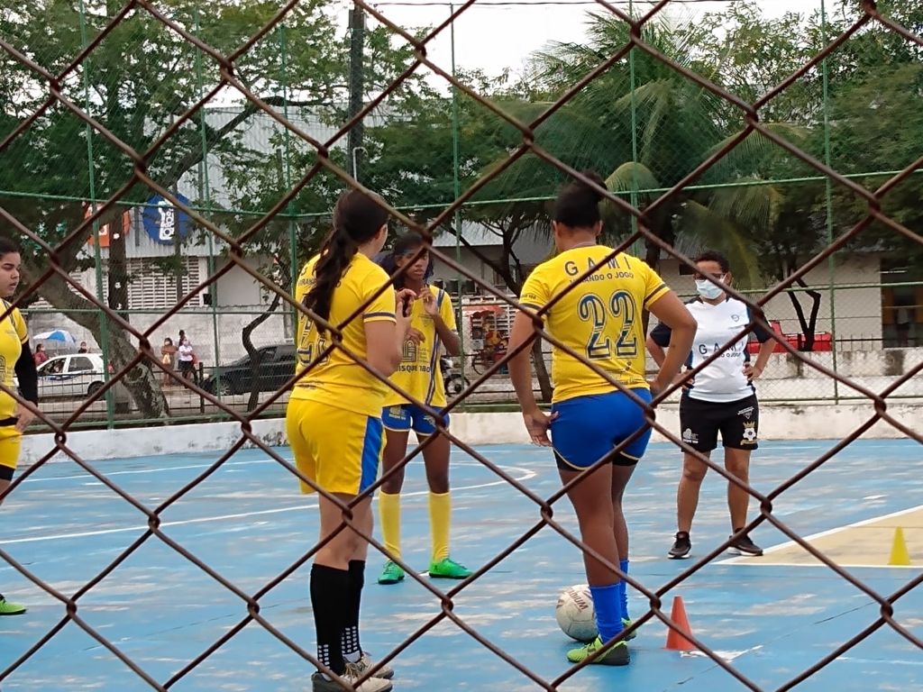 Quatro mulheres jogam uma partida de futebol. Elas vestem uniformes azul e amarelo. A quarta jogadora veste preto e branco.